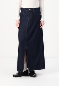 Tummansinisistä denimpitkästä hameesta, jossa on etuhaalari, näkyvät saumat ja vyötärön nappi. Yhdistetty mustiin loafereihin ja vaaleisiin sukkiin.