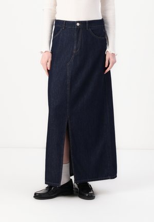Denim maxi skirt in dark blue with a front slit, featuring visible stitching and a waistband button. Paired with black loafers and light socks.