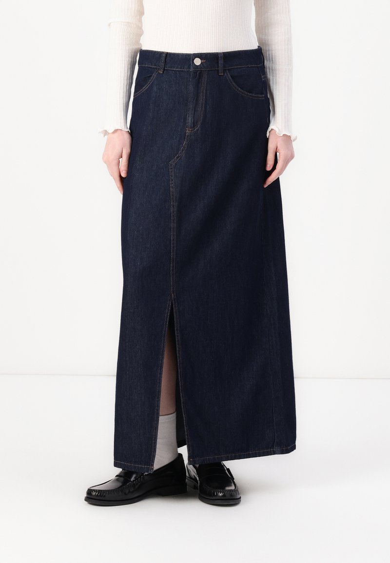 Tummansinisistä denimpitkästä hameesta, jossa on etuhaalari, näkyvät saumat ja vyötärön nappi. Yhdistetty mustiin loafereihin ja vaaleisiin sukkiin.