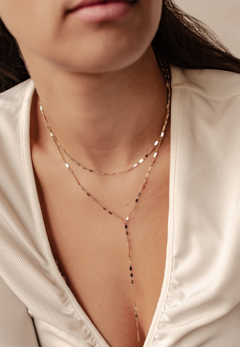 Goldfarbene, mehrlagige Kette mit flachen, rechteckigen Gliedern und einem Y-förmigen Anhänger. Texturierte Oberfläche sorgt für visuelles Interesse. Auf einem hellen Oberteil getragen.