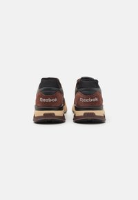 Reebok-sneakers i mörkbrunt mocka och svart nät, med en vadderad tan-färgad sula. Bakifrån syns varumärkesloggan i vitt på hälen.