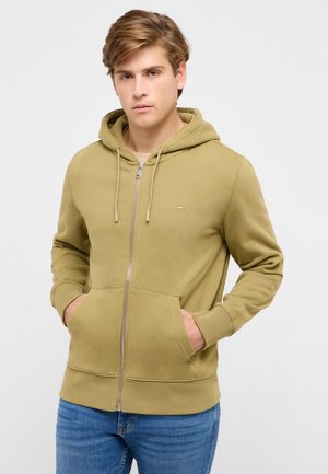 Young man with light brown hair wearing an olive green zip-up hoodie and blue jeans, hands in front pockets, neutral expression.
