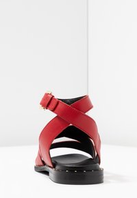 Punaiset nahkakengät, joissa on ristikkäiset hihnat, säädettävä solki ja niittipohja. Rakenne on teksturoitu ylhäältä ja sileä sisältä.