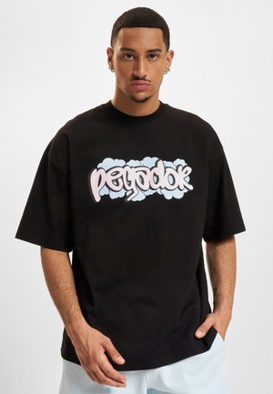 Ung mand med kort hår iført en sort oversized T-shirt med pastelfarvet graffiti tekst og lyse bukser, stående mod en ensfarvet baggrund.