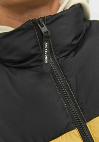 Chaqueta acolchada negra y amarilla con un alto cuello, que presenta una cremallera de longitud completa y costuras en contraste. Nombre de la marca visible en la cremallera.