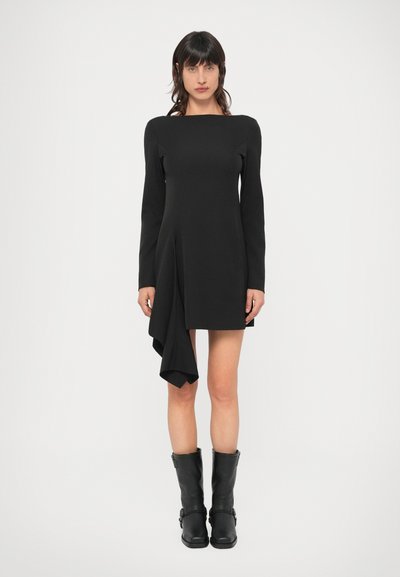 Schwarzes Langarmkleid mit geometrischem Design und asymmetrischem Saum. Kombiniert mit schwarzen kniehohen Stiefeln, präsentiert vor neutralem Hintergrund.