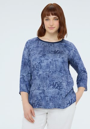 Vrouw met roodbruin haar, draagt hoepeloorbellen, een blauwe lange-mouwen top met patroon en een witte broek, staat met één hand in haar zak.