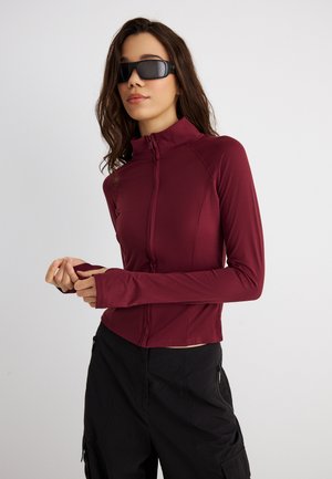 Γυναίκα που φοράει μαύρα γυαλιά ηλίου, μια εφαρμοστή μπουφάν σε μπορντό χρώμα με φερμουάρ και τρύπες για τους αντίχειρες, καθώς και μαύρο παντελόνι cargo, σε απλό ανοιχτό φόντο.