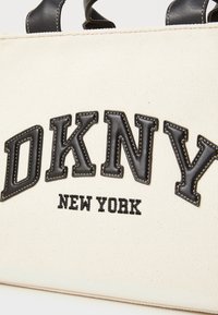 Kreminės spalvos drobinė rankinė su juodais odiniais rankenomis ir nėriniu „DKNY NEW YORK“ ryškiai juoda spalva. Siuvinėtojai pabrėžia dizainą.