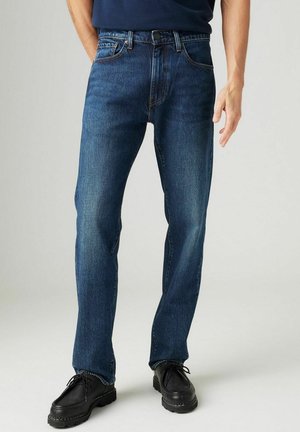 Homme portant un jean droit bleu foncé et des chaussures noires à lacets, debout devant un fond clair uni.