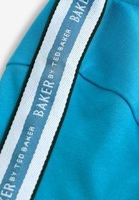 Turkusowy materiał o gładkiej fakturze ma białą i niebieską wyróżniającą się pasek z logo "BAKER by TED BAKER", otoczony ciemnozieloną ramką.