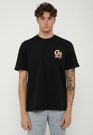 Ung mand med krøllet hår iført en sort T-shirt med teksten "Go for it!" og et smilende ansigt på brystet, samt lysegrå bukser, står stille.