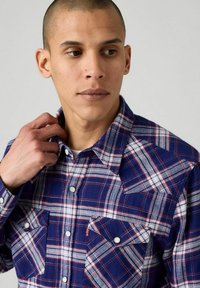 Camicia a quadri blu realizzata in tessuto, con linee bianche e rosse, due tasche sul petto con bottoni a pressione e colletto abbottonato.