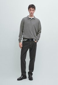 Polo shirt a maniche lunghe grigio sopra una maglietta bianca, abbinato a jeans in denim scuri e mocassini neri. Il modello è in piedi su uno sfondo neutro.