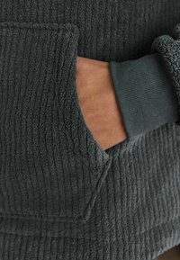 Felpa con cappuccio grigia scura in pile presenta una texture a coste, una tasca a marsupio e un design a contrasto sui polsini. Ingrandimento della mano che si infila nella tasca.