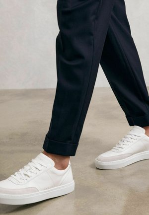 Biele nízke tenisky s textúrovanými detailmi, plochými šnúrkami a miernym polstrovaním; nosené s tmavomodrými šitými nohavicami s manžetami.