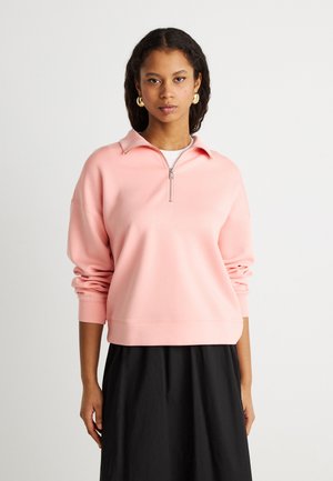 Giovane donna con felpa rosa con mezza cerniera e maniche lunghe, indossando una gonna nera, in piedi davanti a uno sfondo chiaro uniforme.