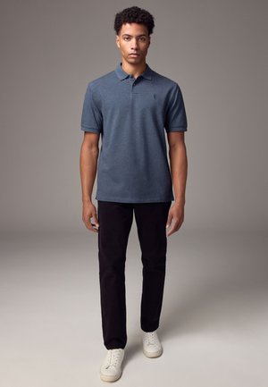 Moški stoji, oblečen v modro polo majico s kratkimi rokavi, črne hlače in bele superge, na enobarvnem sivem ozadju.