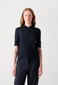 Cămașă polo cu mânecă scurtă de culoare bleumarin, realizată dintr-un material tricotat moale. Prezintă un guler și o deschidere cu cinci butoane. Asortată cu pantaloni închiși la culoare.