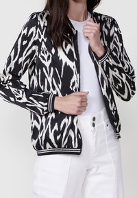 Femme portant une veste bomber à motif abstrait noir et blanc sur une chemise blanche et un pantalon blanc taille haute avec deux boutons.