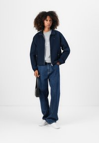 Navyblauwe zip-up jack met een relaxte pasvorm, gecombineerd met een lichtgrijze sweater en wijdvallende blauwe jeans. Model houdt een zwarte schoudertas vast.