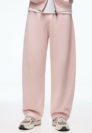 Person iført løse lyserøde sweatpants, hvide sneakers og en delvist lynet matchende lyserød jakke.