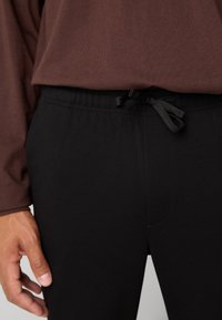 Primer plano de una persona que lleva pantalones negros con un cordón negro atado y una camisa de manga larga marrón, con la mano descansando a su lado.