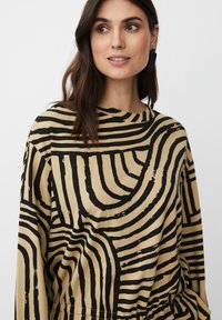 Maglietta a maniche lunghe in tessuto beige con motivi a onde neri, caratterizzata da un collo rotondo e una vestibilità rilassata. Materiale leggero e texturizzato.