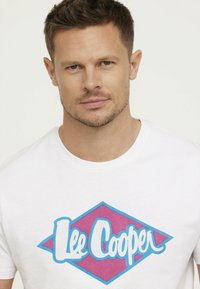 T-shirt en coton blanc avec un grand symbole de diamant rose aux contours bleus et le texte "Lee Cooper" en blanc, centré sur le devant.