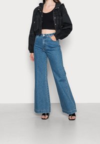 Schwarze, cropped Jacke mit Kapuze, kombiniert mit hoch taillierten, weiten blauen Jeans. Schwarze Riemchensandalen runden das Outfit ab.