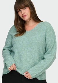 Šviesiai žalias V formos kaklo megztinis su rištu raštais ir ilgomis rankovėmis. Audinys atrodo minkštas ir šiek tiek laisvas, turintis atsipalaidavusį stilių.