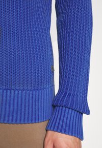 Pull bleu à côtes avec un tissu texturé, des poignets contrastants et une étiquette logo discrète à l'ourlet. Gros plan sur les détails de la manche.