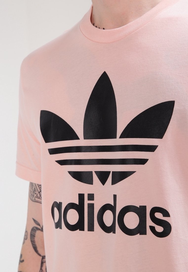 T-shirt en coton rose avec un grand logo Adidas trefoil noir et du texte. Manches courtes avec un col rond. Texture lisse.
