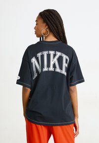Black cotton t-shirt with short sleeves, featuring large, silver "NIKE" text on the back. Contrast stitching adds subtle detail. Paired with orange pants.