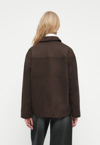 Bruine faux suède jas met een eenvoudige kraag, verlaagde schouders en een relaxed fit. Gelaagde textuur met strakke lijnen en een rugnaad detail.