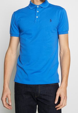Homme portant un polo près du corps de couleur bleu vif à manches courtes, avec trois boutons blancs et un petit logo foncé sur la poitrine.