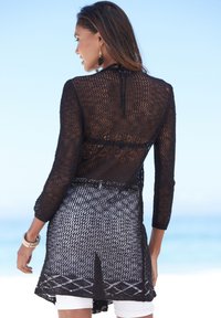 Cardigan nero a maglia aperta con maniche lunghe, caratterizzato da un design in pizzo e un orlo spaccato. Indossato sopra pantaloni corti bianchi, che mettono in risalto un motivo dettagliato.