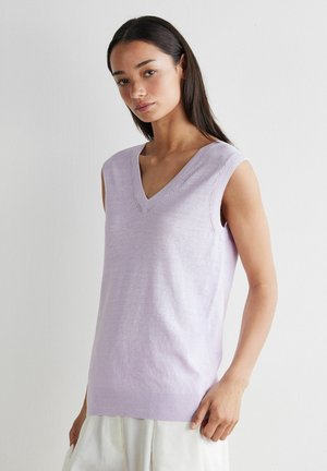 Jeune femme aux longs cheveux foncés portant un gilet sans manches lavande à col en V et un pantalon de couleur claire, sur fond blanc uni.