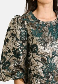Frau trägt ein metallisch goldenes und grünes Blumenbrokat-Oberteil mit gepufften Ärmeln und rundem Halsausschnitt.