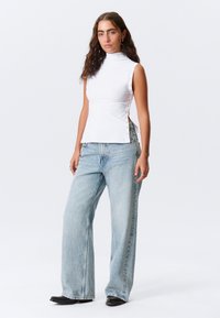 Λευκή μπλούζα με ψηλό λαιμό και λεπτομέρειες με κουμπιά στο πλάι, συνδυασμένη με ανοιχτό μπλε τζιν παντελόνι φαρδύ και μαύρες μπότες με μυτερό δάχτυλο, σε απλό φόντο.