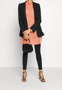 Femme portant un blazer noir par-dessus une robe florale corail avec des leggings noirs, tenant un sac à main noir et portant des talons hauts noirs à lanières.