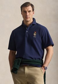 Άνδρας που φοράει μπλε σκούρο πόλο πουκάμισο με κεντημένο λογότυπο, χακί παντελόνι, πράσινο πουλόβερ δεμένο στη μέση και ρολόι χειρός, στέκεται μπροστά σε ουδέτερο φόντο.