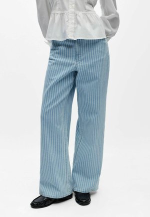 Frau trägt hellblaue Jeans mit weitem Bein und weißen vertikalen Streifen, kombiniert mit schwarzen flachen Schuhen und einer weißen langärmligen Bluse.