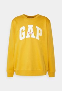 Sweat-shirt jaune avec col rond, manches longues et poignets côtelés. Présente un grand logo "GAP" blanc avec "Original" en rouge en dessous. Le matériau a une texture lisse.