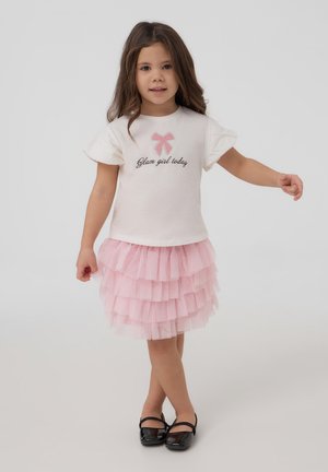 T-shirt bianca in cotone con maniche a volant e la scritta "Glam girl today", abbinata a una gonna in tulle rosa a strati. Le scarpe nere completano l'outfit.