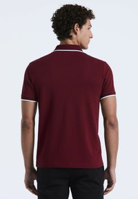 Mann mit lockigem Haar, der ein kurzärmeliges bordeauxrotes Poloshirt mit weißen Details trägt, von hinten vor einem schlichten hellen Hintergrund gesehen.