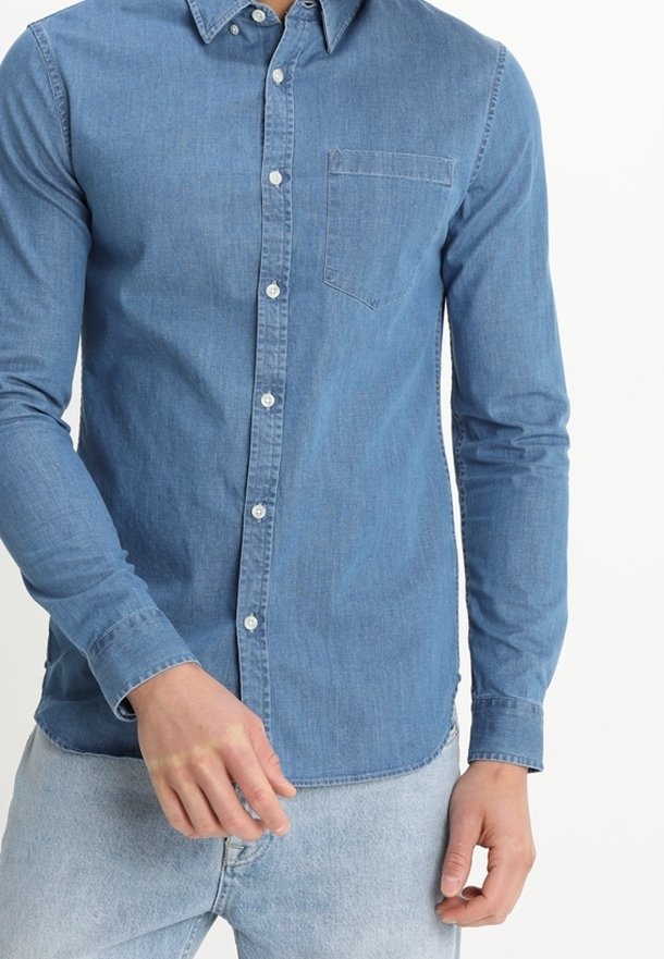 Homme portant une chemise en jean bleu clair à boutons avec manches longues et un jean bleu clair, debout devant un fond uni.