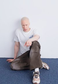 White t-shirt with small patch, loose olive green pants, and beige sneakers with patterned accents. Person seated against a white background.