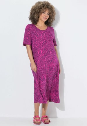 Mujer con cabello rizado que lleva un vestido midi rosa con estampado de hojas y sandalias rosas a juego, de pie sobre un fondo blanco.