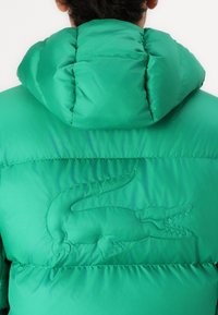 Zelena puffer jakna s prešanim dizajnom, visokim kragnom i ukrasnim vezom krokodila na leđima.
